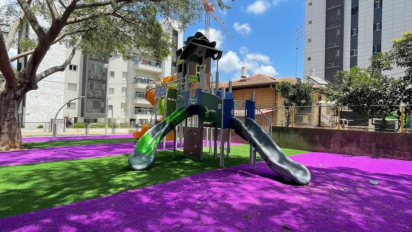 ריצוף גומי בטיחותי בגווני סגול וירוק במתקן משחקים סמוך למבני מגורים