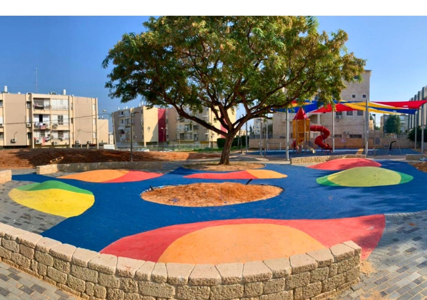ריצוף גומי צבעוני בגן ציבורי סביב עץ, עם מתקן משחק צבעוני ברקע